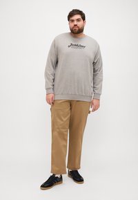 Sweat-shirt gris avec des lettres noires, associé à un pantalon beige et des baskets noires. Coupe décontractée avec des manches longues et des poignets côtelés.