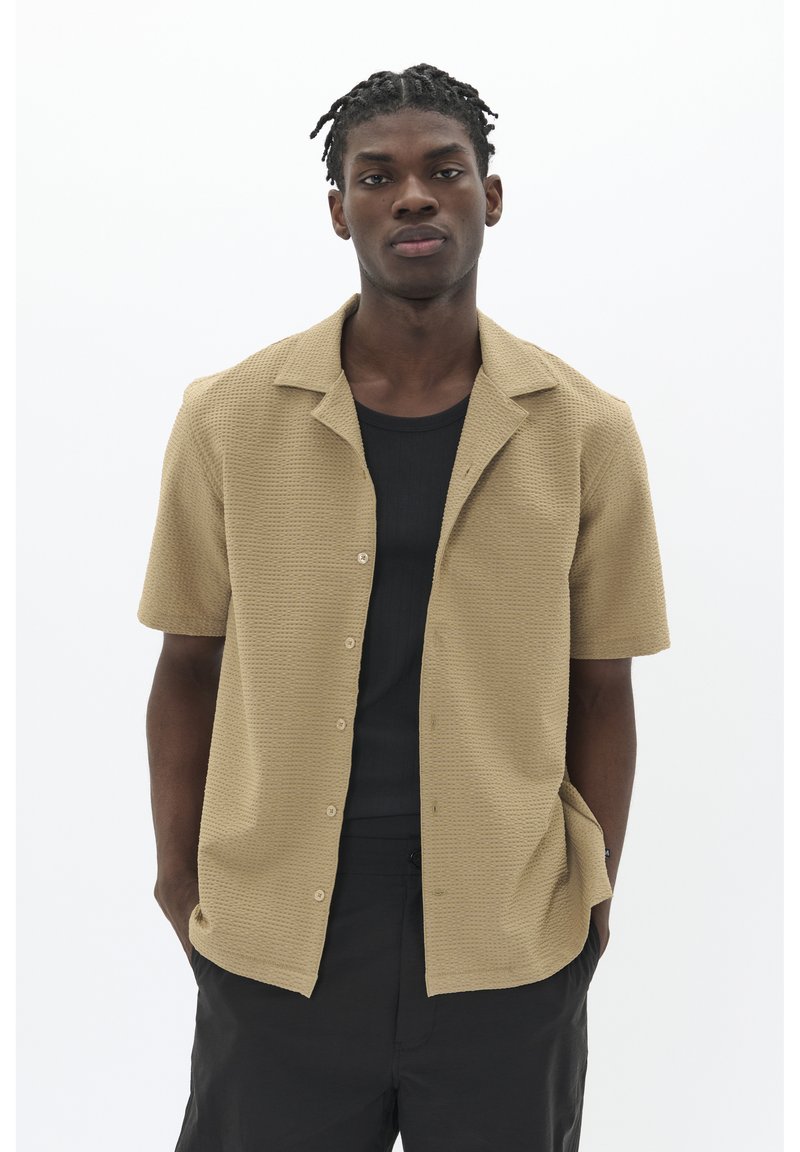 Camisa beige de manga corta hecha de tela texturizada con una parte delantera abotonada, combinada con una camiseta negra y pantalones oscuros.