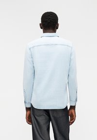 Camisa de ganga azul claro com mangas compridas, punhos com botões e colarinho pontiagudo. Apresenta uma bainha curva e um padrão desbotado.