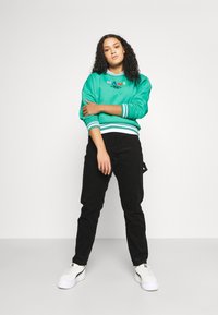 Zelena dukserica s bijelim i tamnoplavim prugama na manžetama i rubu, s šarenim natpisom "KANI"; uparena s crnim hlačama i bijelim tenisicama.