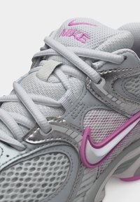 Belo-siva tekaška copata Nike z rožnatimi poudarki, mrežasto tkanino, natančnim šivanjem in belimi vezalkami, ki se križajo čez jeziček z logotipom Nike na vrhu.