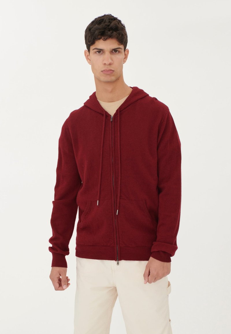 Röd zip-up hoodie i texturerat material, med huva, dragband och framfickor. Modellen bär ljusa byxor.