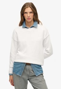 Jonge vrouw in een witte trui over een spijkerblouse en gestreepte broek, staande voor een effen witte achtergrond.