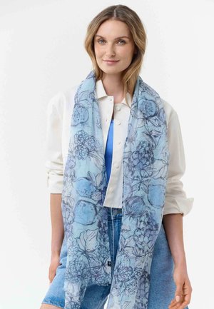 Woman wearing a white jacket and blue top, draped with a light blue floral-patterned scarf, standing against a white background.