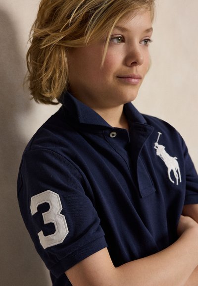 Vaaleahiuksinen lapsi laivastonsininen pikeepaita, jossa hihassa numero 3 ja rinnassa hevospelaajan logo, kädet ristissä, katsoo oikealle.