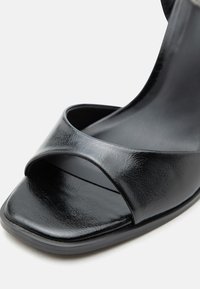 Μαύρο σανδάλι με ανοιχτά δάχτυλα, φαρδιά λουράκι μπροστά και τετράγωνη μύτη, που δείχνει λεία, ελαφρώς γυαλιστερή δερμάτινη υφή.