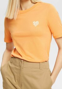 Orange bomull t-shirt med ett vitt hjärtgrafik och texten "ALL WE NEED IS NAPS." Korta ärmar, rundad halsringning, slät textur.