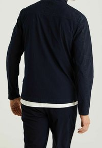 Navy blue jacket in lightweight fabric with long sleeves, featuring a straight cut and minimal stitching details. Layered over a white top.