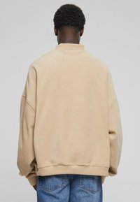 Beige oversized genser med senkede skuldre, ribbestrikkede mansjetter og klassisk rund hals, laget av mykt stoff. Bakside viser.