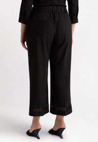 Svarta korta byxor med elastisk midja, som har utskuren spetskant vid fållen och två bakfickor. Materialet verkar lätt och mjukt.