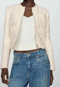 Lys pink, tekstureret jakke med fire frontlommer, rund halsudskæring og synlige sømme, parret med en hvid tanktop og blå denim jeans.