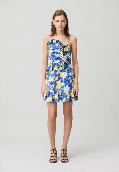 La robe florale présente un fond bleu avec des fleurs jaunes et blanches, un décolleté et un ourlet à volants, ainsi que des bretelles fines. Associée à des sandales noires à lanières.