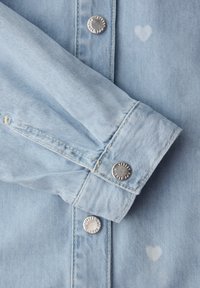 Manche en denim bleu clair avec boutons-pression en métal et motifs de petits cœurs blancs discrets sur le tissu près du poignet et de la patte de boutonnage.