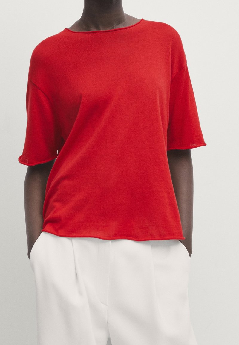 Personne portant un haut rouge vif à manches courtes rentré dans un pantalon blanc taille haute, les mains dans les poches, debout devant un fond uni.