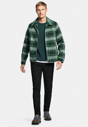 Green plaid zip-up jacket with a collar, worn over a dark green sweater, paired with black pants and brown hiking boots.