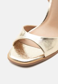Χρυσό μεταλλικό σανδάλι με ανοιχτά δάχτυλα, επίπεδη σόλα και φαρδιά λωρίδα πάνω από τα δάχτυλα, που δείχνει υλικό με υφή και γυαλιστερή επιφάνεια.