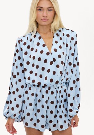 Femme blonde portant une combinaison bleu clair à manches longues avec de gros pois marron foncé et une ceinture assortie à la taille.