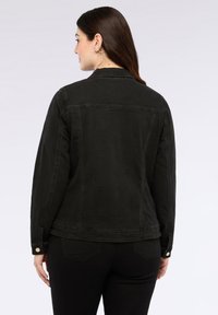 Nainen, jolla on pitkät ruskeat hiukset, suuret rengaskorvakorut, musta denimpussukka ja mustat housut, selkä kameraan päin, yksinkertaista taustaa vasten.