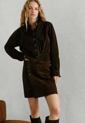 Kobieta o długich blond włosach, ubrana w ciemnobrązową, zapinaną na guziki marynarkę oraz dopasowaną spódnicę, stojąca na tle jasnej, teksturowanej ściany.