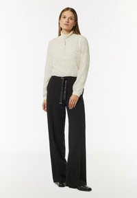 Hellbeige Langarmtop mit strukturiertem Muster und halbem Reißverschluss, kombiniert mit hochgeschnittenen schwarzen weit geschnittenen Hosen und schwarzen Schuhen.