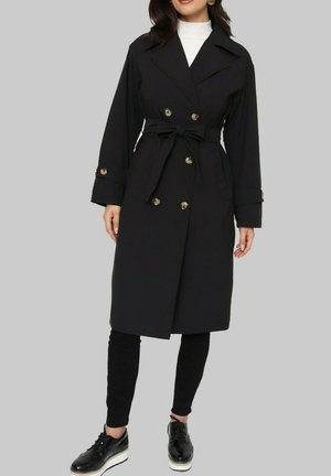 Manteau noir à double boutonnage, avec des boutons dorés, une taille ceinturée et de longues manches. Porté sur un pantalon noir et des chaussures.