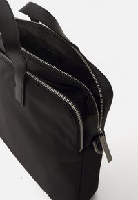 Bolso tote de nailon negro con cremalleras metálicas dobles, acentos de cuero y asas texturizadas. El interior cuenta con un forro suave.
