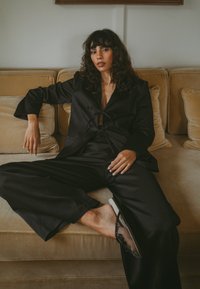 Črn krojen suknjič z širokimi hlačami in poudarjenim pasom, v kombinaciji s črnimi japonkami z okrasnimi detajli, postavljenimi ob bež kavču.