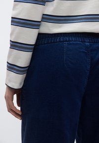 Pantalones vaqueros azules con una textura suave, que cuentan con una cintura elástica y un bolsillo trasero, combinados con una camisa de manga larga blanca con rayas.