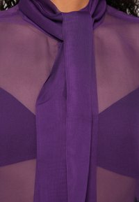 Paarse blouse met doorschijnende stof en een gladde textuur, voorzien van een strikkraagdetail en gelaagde designelementen.