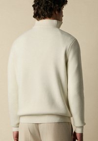 Světlý béžový svetr s vysokým límcem, ribovaný vzor a přiléhavé manžety. Hladká tkanina s jemným leskem, nošena přes béžové kalhoty.