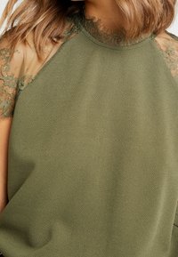 Mujer con una blusa verde oliva que tiene mangas de encaje con flores y un detalle de collar de encaje, mostrada de los hombros a la cintura.