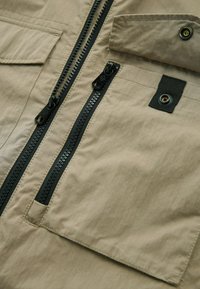 Jachetă de exterior bej cu două fermoare negre, material texturat, un buzunar tip patch și detalii cu nasturi lângă guler.