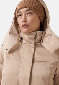 Beige wasserfeste Jacke mit hohem Kragen und Druckknopfdetails, mit einer glatten, glänzenden Textur. Kombiniert mit einer gerippten braunen Strickmütze.