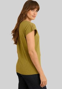 Moteris su ilgais rudais plaukais, dėvinti alyvuogių žalios spalvos trumpomis rankovėmis marškinėlius su skylėmis ant pečių ir tamsias kelnes, žiūri per petį.