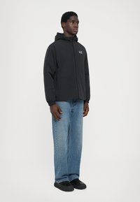 Svart zip-up jacka med huva, med logotyp på bröstet; matchas med ljusblå vida jeans och svarta slip-on skor.