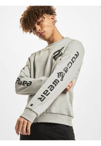 Grå sweatshirt lavet af blødt materiale, med sort trykt tekst på ærmerne og et fremtrædende logo på brystet. Tætsiddende manchetter og kant.