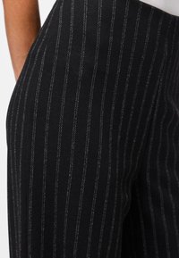 Lähikuva henkilöstä, jolla on päällään mustat housut, joissa on ohuet valkoiset pystysuorat raidoitukset, ja valkoinen paita, joka on sullottu housuihin.