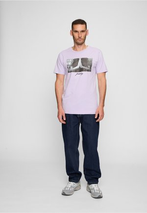 Mann steht und trägt ein lavendelfarbenes T-Shirt mit einer schwarz-weißen Grafik betender Hände und dunkelblaue Jeans vor einem schlichten Hintergrund.