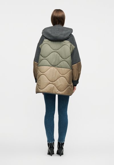 Γυναίκα που φοράει γκρι, πράσινο ελιάς και καμηλό μπουφάν με κουκούλα και σχέδιο από διαφορετικά χρώματα, με στενά μπλε τζιν και μαύρα μποτάκια με τακούνια.