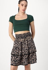 Zöld, rövid ujjú crop top, amelyet egy fekete és virágmintás, réteges szoknyával párosítottak, smockolt derékkal és díszes aranyszínű övvel.