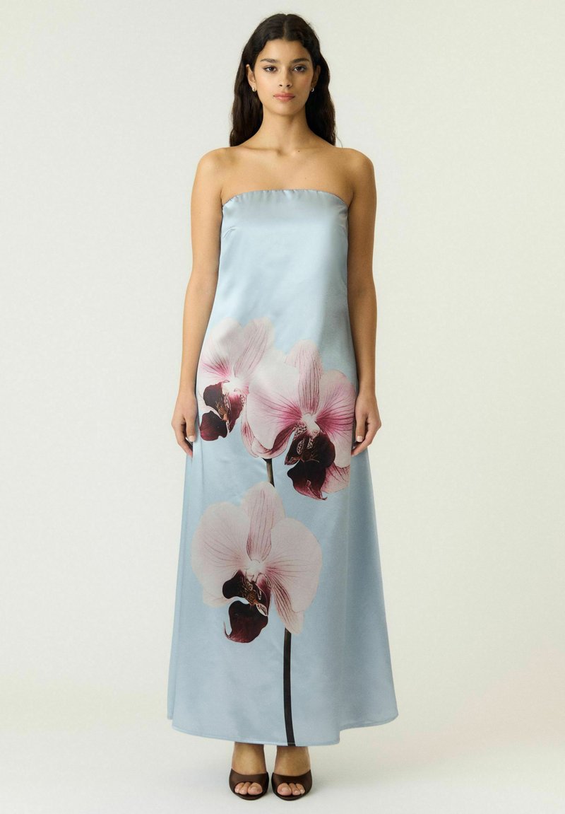 Femme portant une robe bustier en satin bleu clair avec de grandes impressions d'orchidées roses, debout, les bras détendus le long du corps.