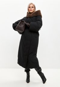 Sort lang dunjakke med en brun pelskrave, der har en indsnøret talje, store ærmer og spidse sorte stiletthæle. Brun håndtaske som accent.