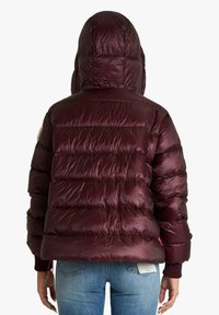 Persona che indossa una giacca imbottita lucida color bordeaux con cappuccio, abbinata a jeans blu, vista da dietro contro uno sfondo bianco.