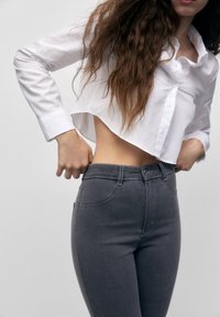 Femme aux longs cheveux ondulés portant un t-shirt blanc court ajustant un jean skinny taille haute gris foncé sur un fond clair uni.