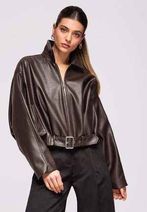 Jeune femme avec une queue de cheval lisse portant une veste en cuir marron foncé ceinturée et un pantalon noir, debout devant un fond clair uni.