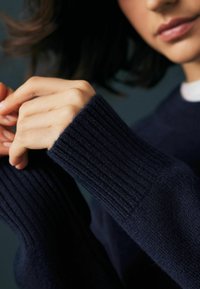 Temno modra pulover s rebrastimi manšetami in mehkimi teksturami; roke so sklenjene, naravni nohti so vidni, v ozadju je zamegljena slika.