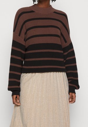 Personne portant un pull rayé marron et noir à manches longues et une jupe plissée beige clair sur un fond uni.