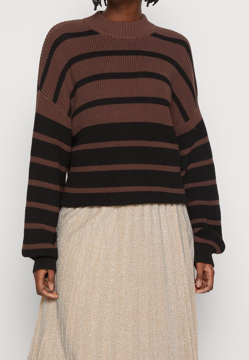 Personne portant un pull rayé marron et noir à manches longues et une jupe plissée beige clair sur un fond uni.