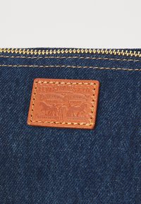 Denimveske med en teksturert lys brun lærpatch med preget design, gullglidelås og kontrasterende gule sømmer på mørkt stoff.