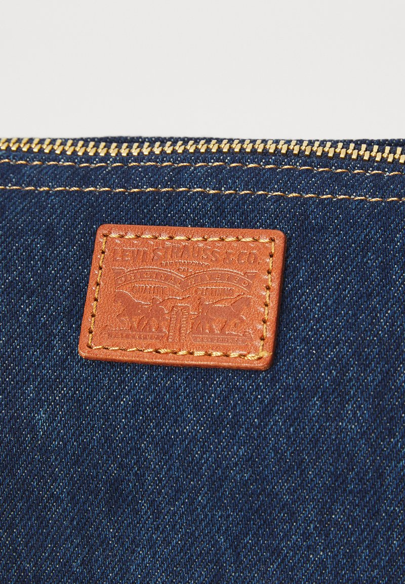 Bolsa de denim con un parche de cuero texturizado en color marrón claro, que presenta un diseño en relieve, cierre dorado y costuras amarillas que contrasta sobre la tela oscura.
